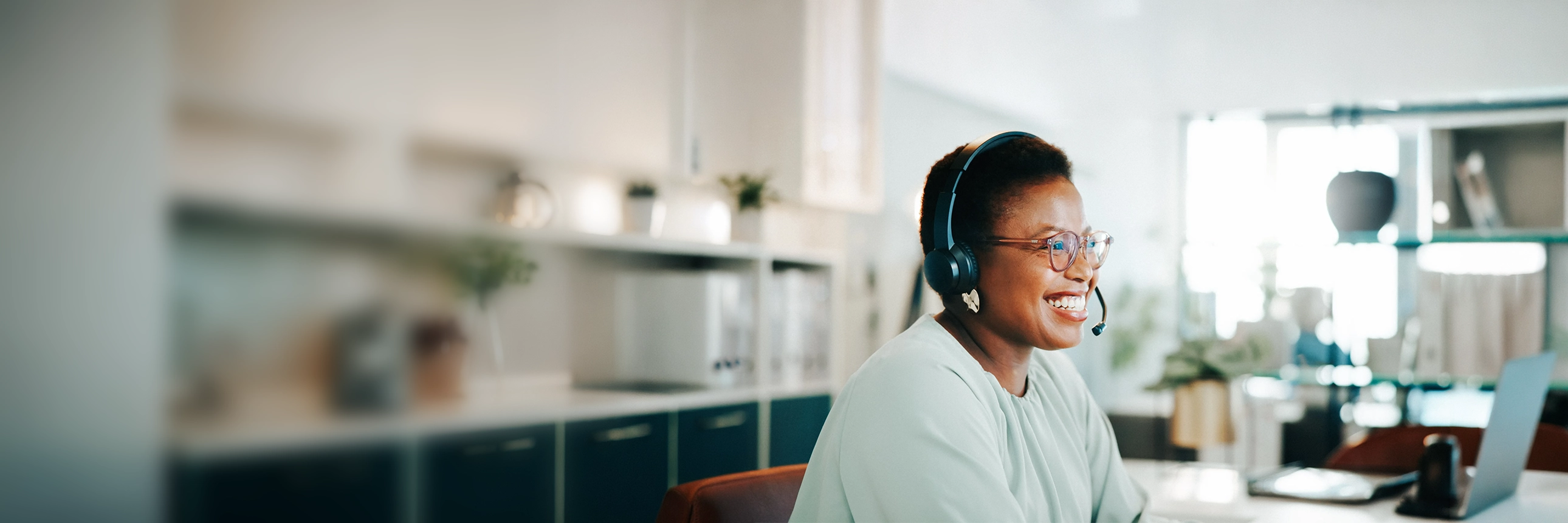 Person wearing a headset seated at a desk in an office, with a computer and shelves in the background. 