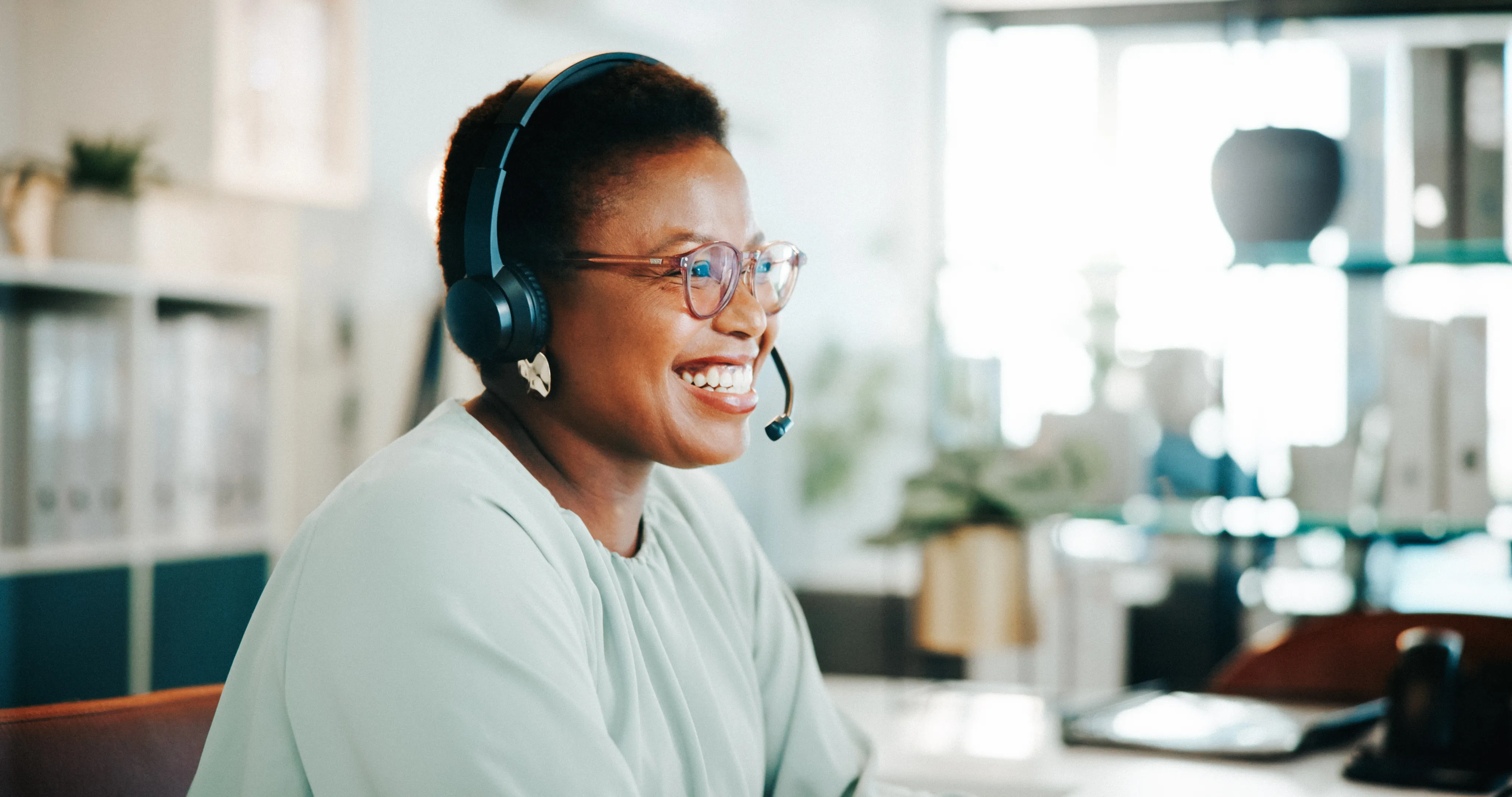 Person wearing a headset seated at a desk in an office, with a computer and shelves in the background. 