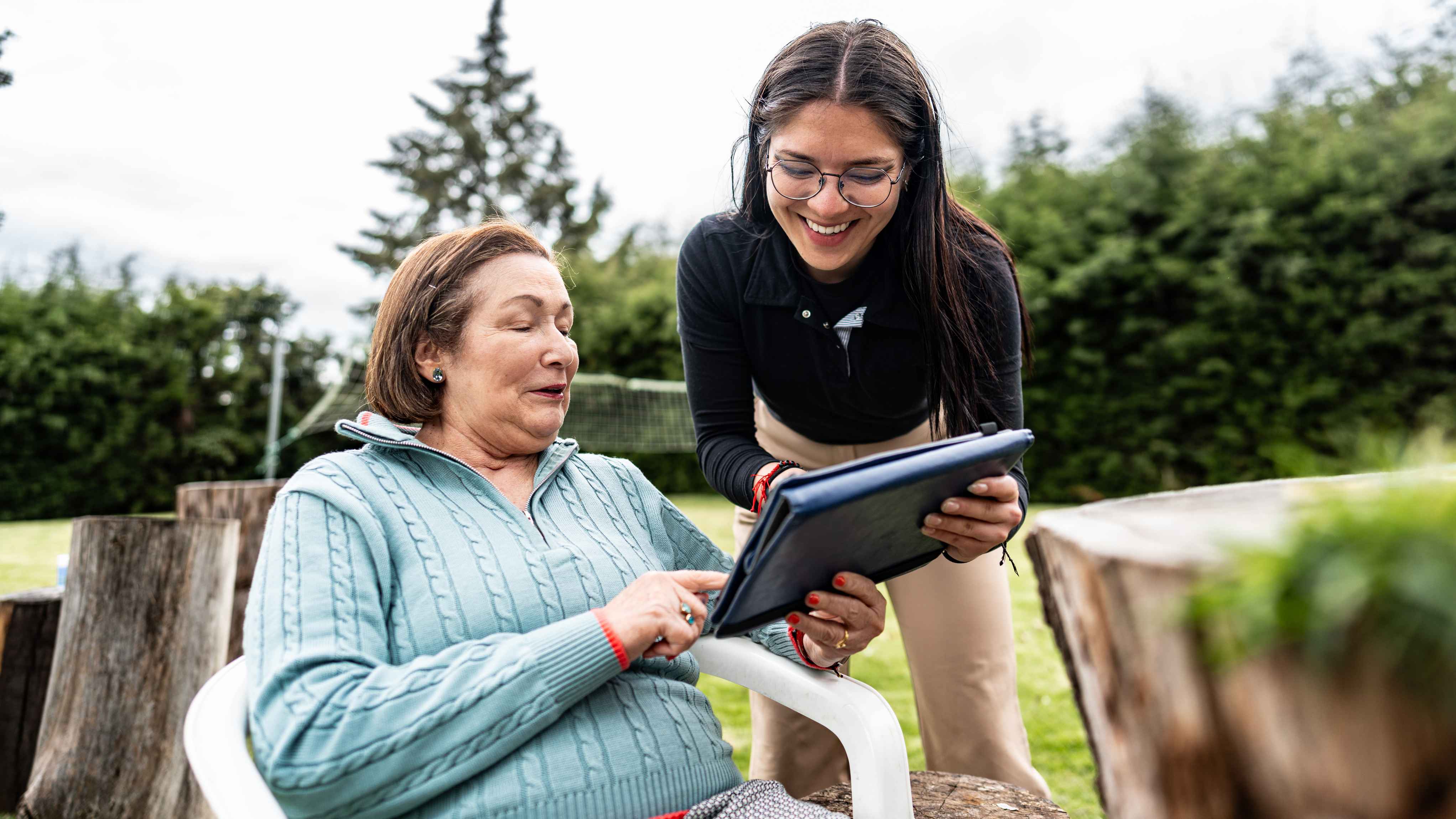 An older woman sits outside on a white plastic chair, smiling as she interacts with a digital tablet held by a young female carer standing beside her. The younger woman is wearing glasses and a black top, and both appear to be enjoying the moment. The background features green trees, a lawn, and a net, suggesting a relaxed garden or park setting.
