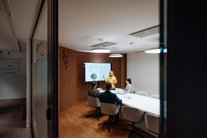 Woman presenting data charts on a screen to three colleagues seated around a conference table in a modern meeting room.