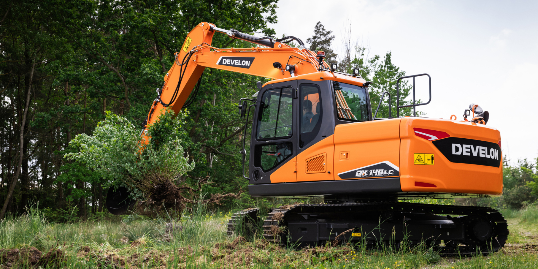 A DEVELON crawler excavator moving large brush and limbs on a landscaping job site.