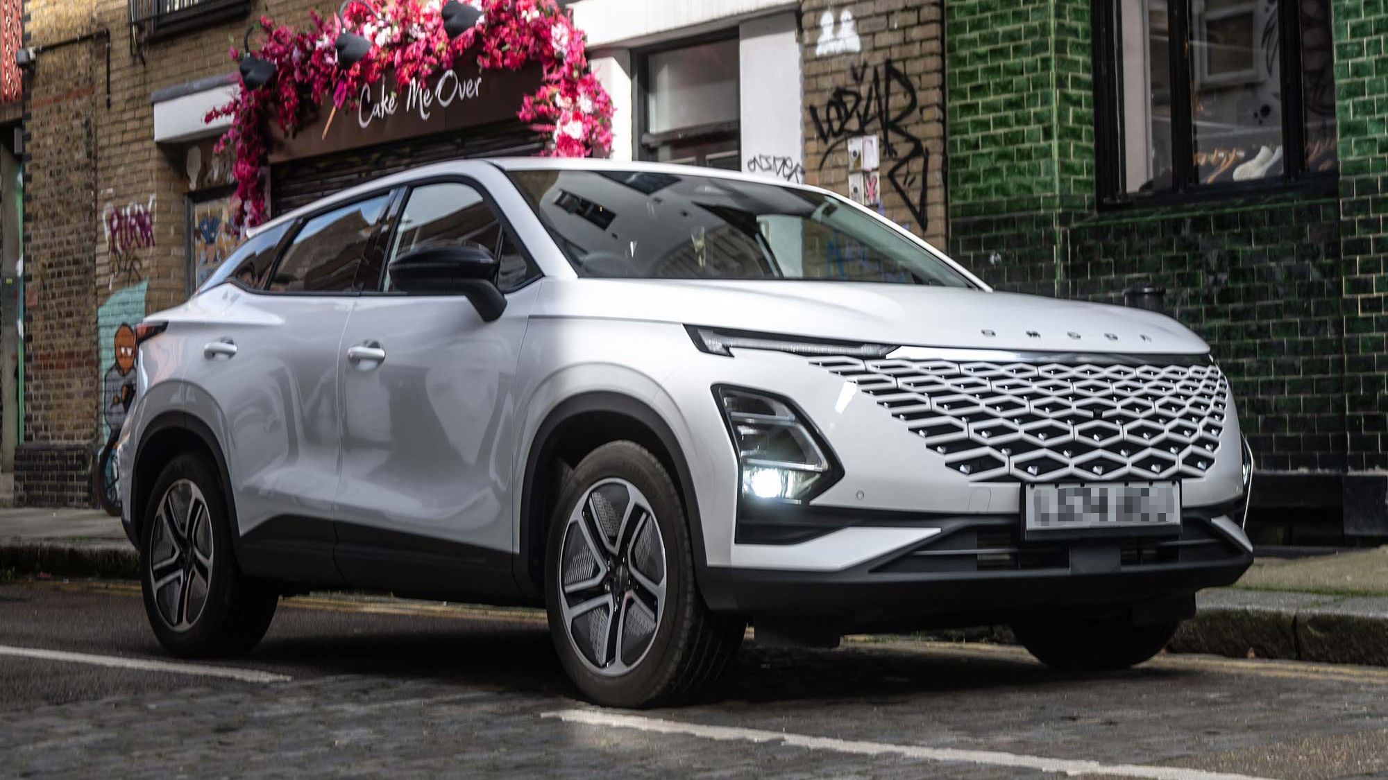 A white Omoda 5 SUV parked on a cobbled city street, featuring a futuristic grille design and angular LED lights.