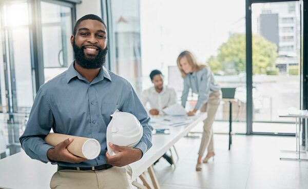Telecommunications project lead standing in an office holding architectural plans and a hard hat with colleagues in the background.