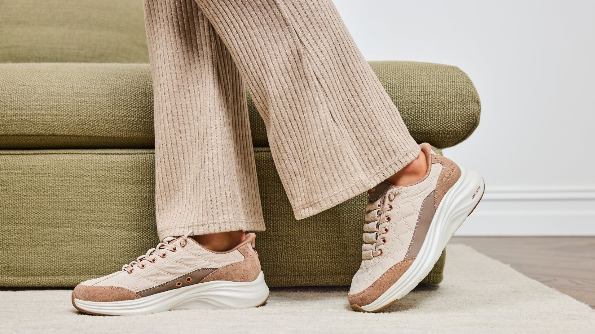 Person seated on a lounge wearing beige Skechers Cozy Fit sneakers with a grey knit jumper and ribbed pants.