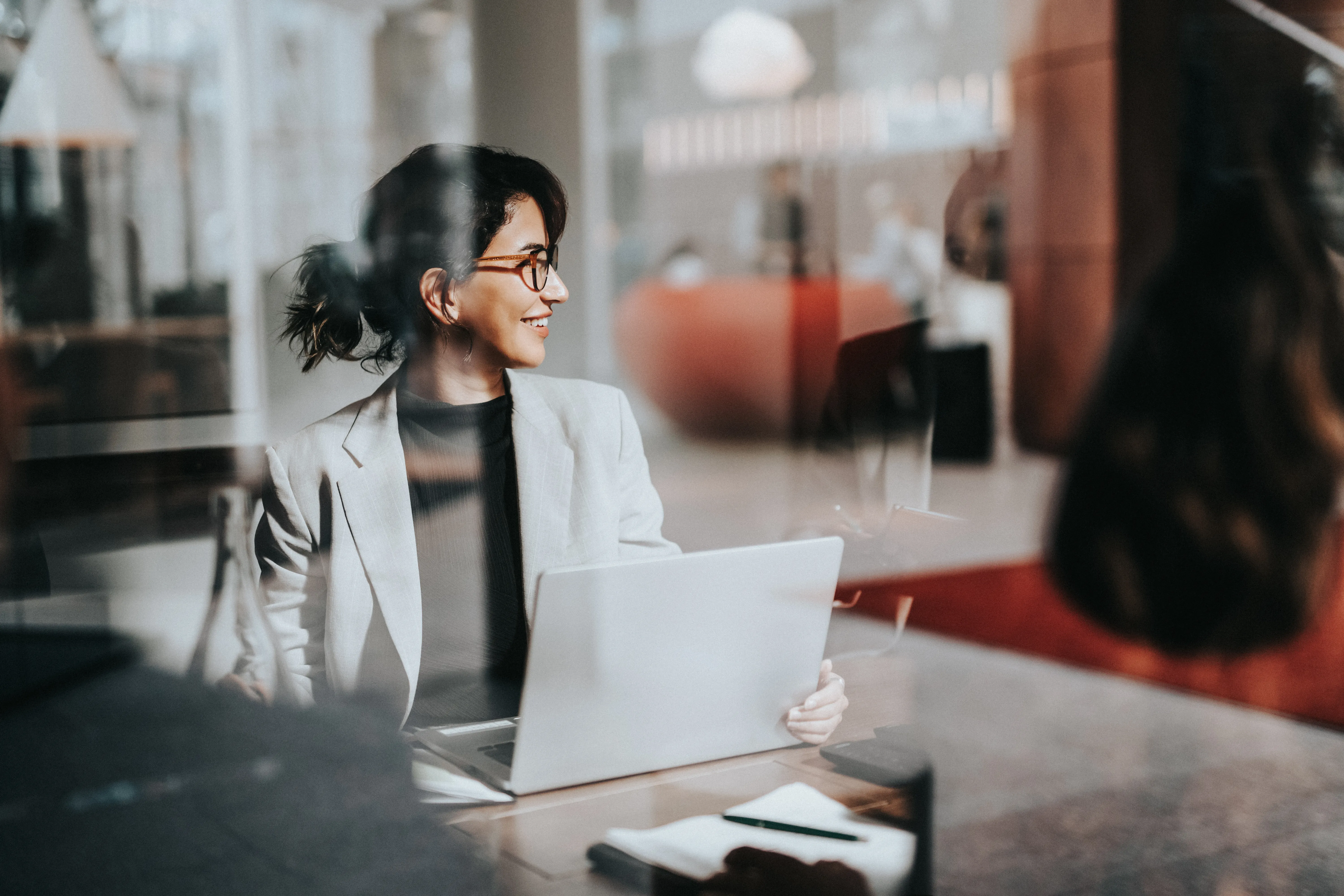 Smiling business woman in office on laptop seen through glass window 