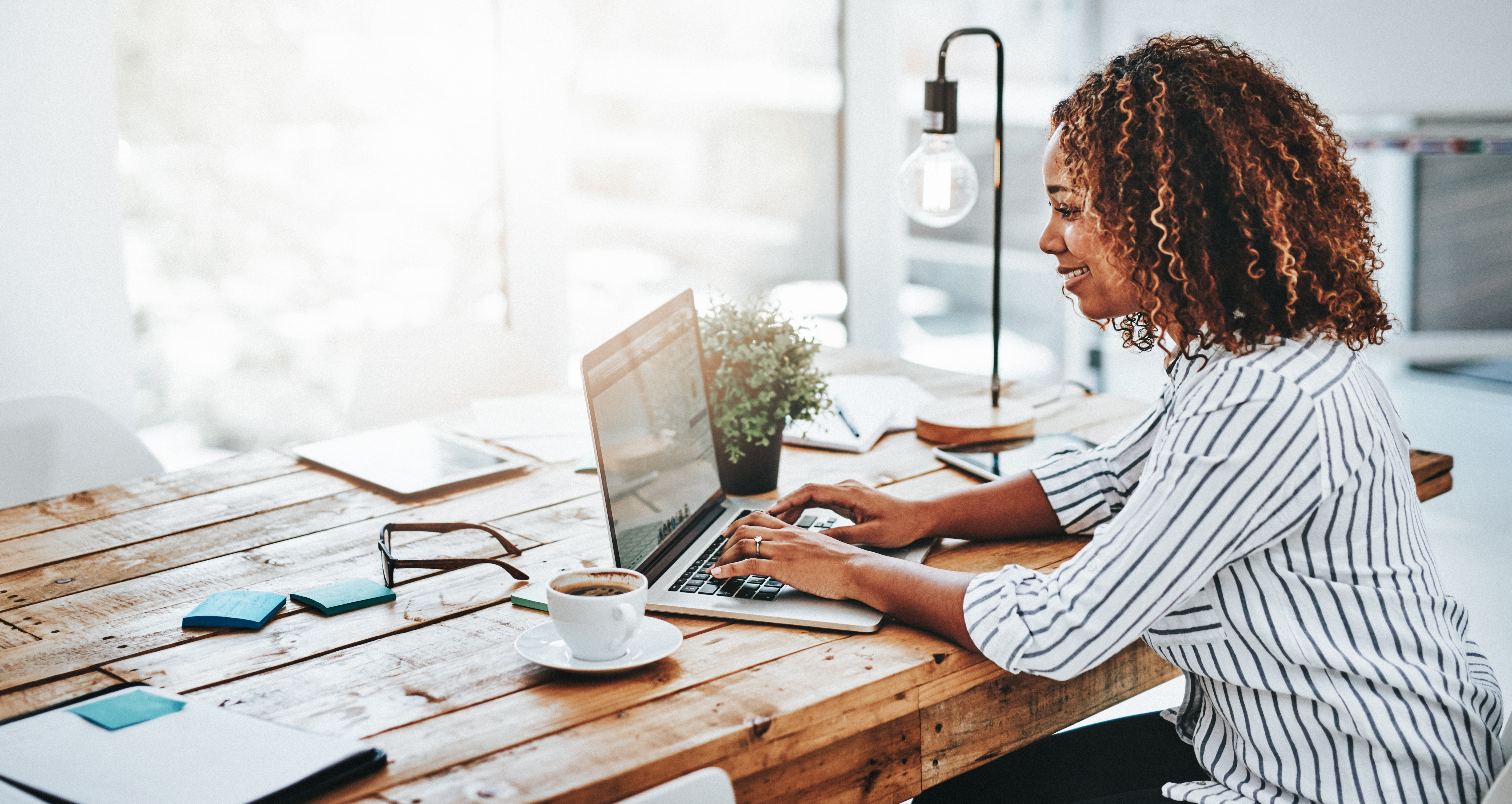 Professional African American woman sitting at her desk in brightly lit office working on laptop 