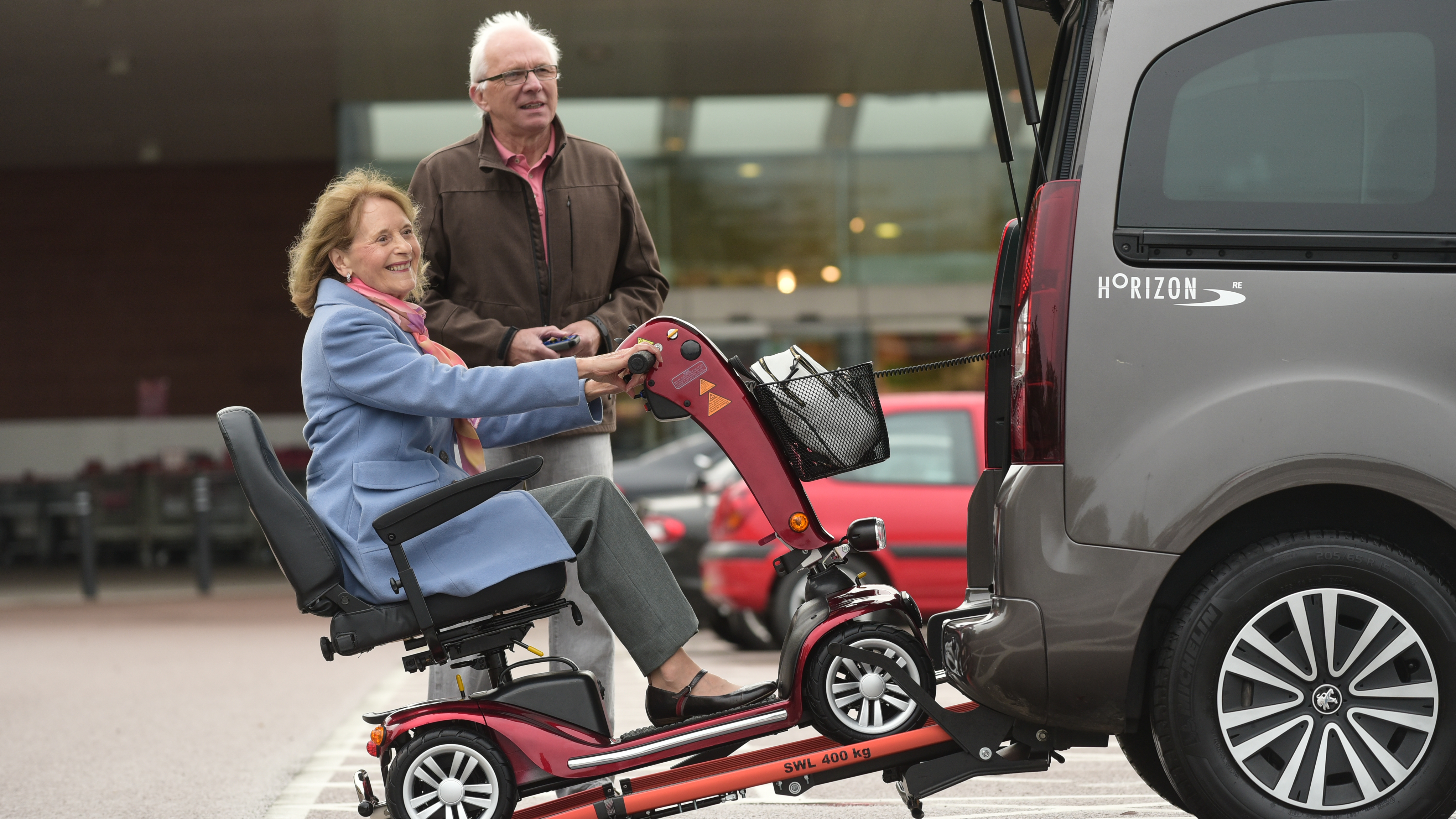 An older woman smiles as she drives her red mobility scooter up a rear ramp into a grey Horizon WAV (wheelchair accessible vehicle), while an older man stands nearby with the remote control.