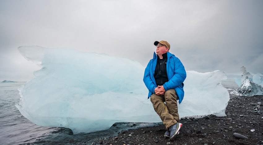 A man in a blue coat sits on a block of blue ice on the coast of Iceland