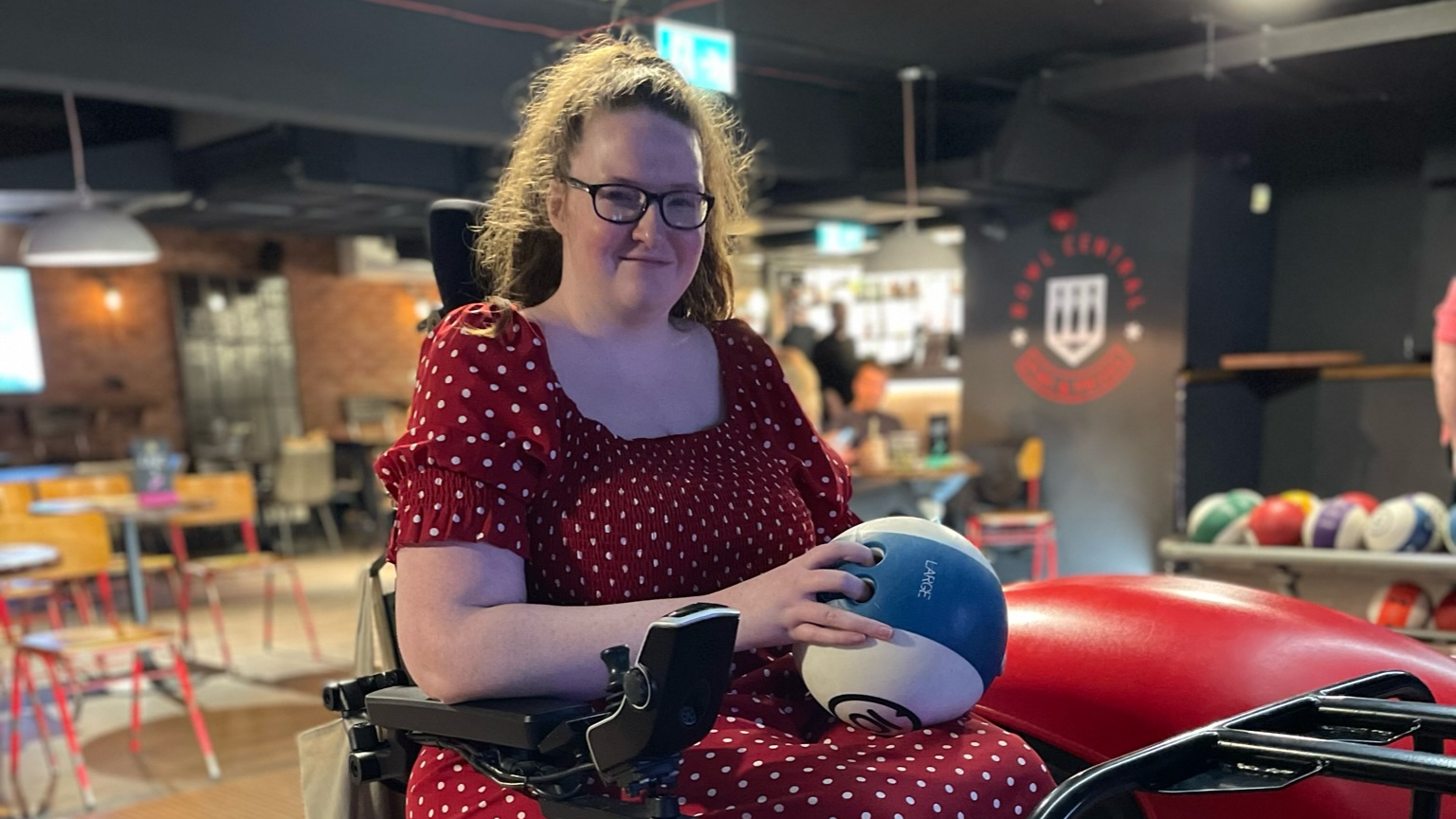 Shona Louise, wearing a red polka dot dress, smiles while seated in a power wheelchair at a bowling alley. She holds a blue and white bowling ball, ready to play, with colourful bowling balls visible in the background.