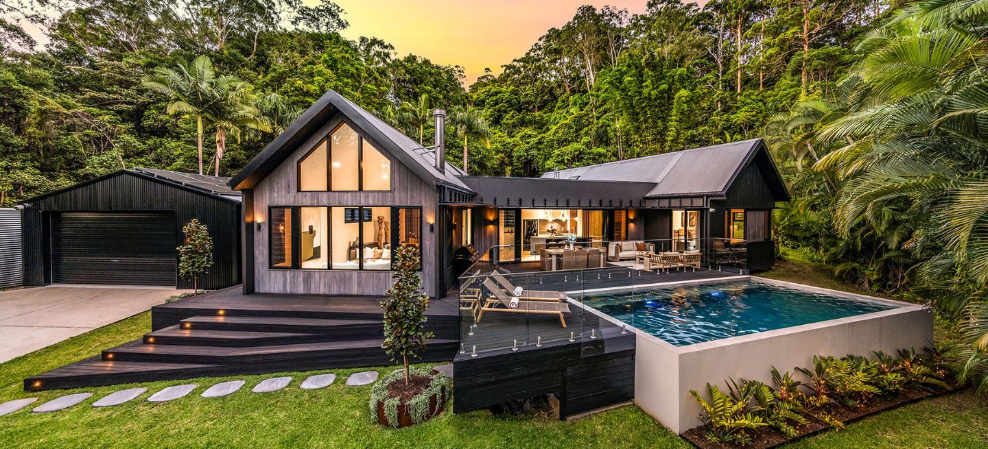 yourtown Prize Home Draw 545. Dusk external image from the front of a single-level pavilion-style house with black cladding. There is an alfresco area at the front with a pool. A garage and driveway is on the left hand side. Lush gardens surround the home.