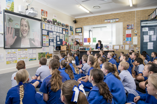 A group of school children sit on the floor and stare up at a projector screen. On the screen, a Kids Helpline Counsellor waves back.