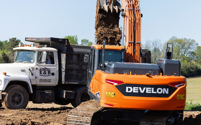 A DEVELON excavator empties dirt from a bucket into a dump truck.