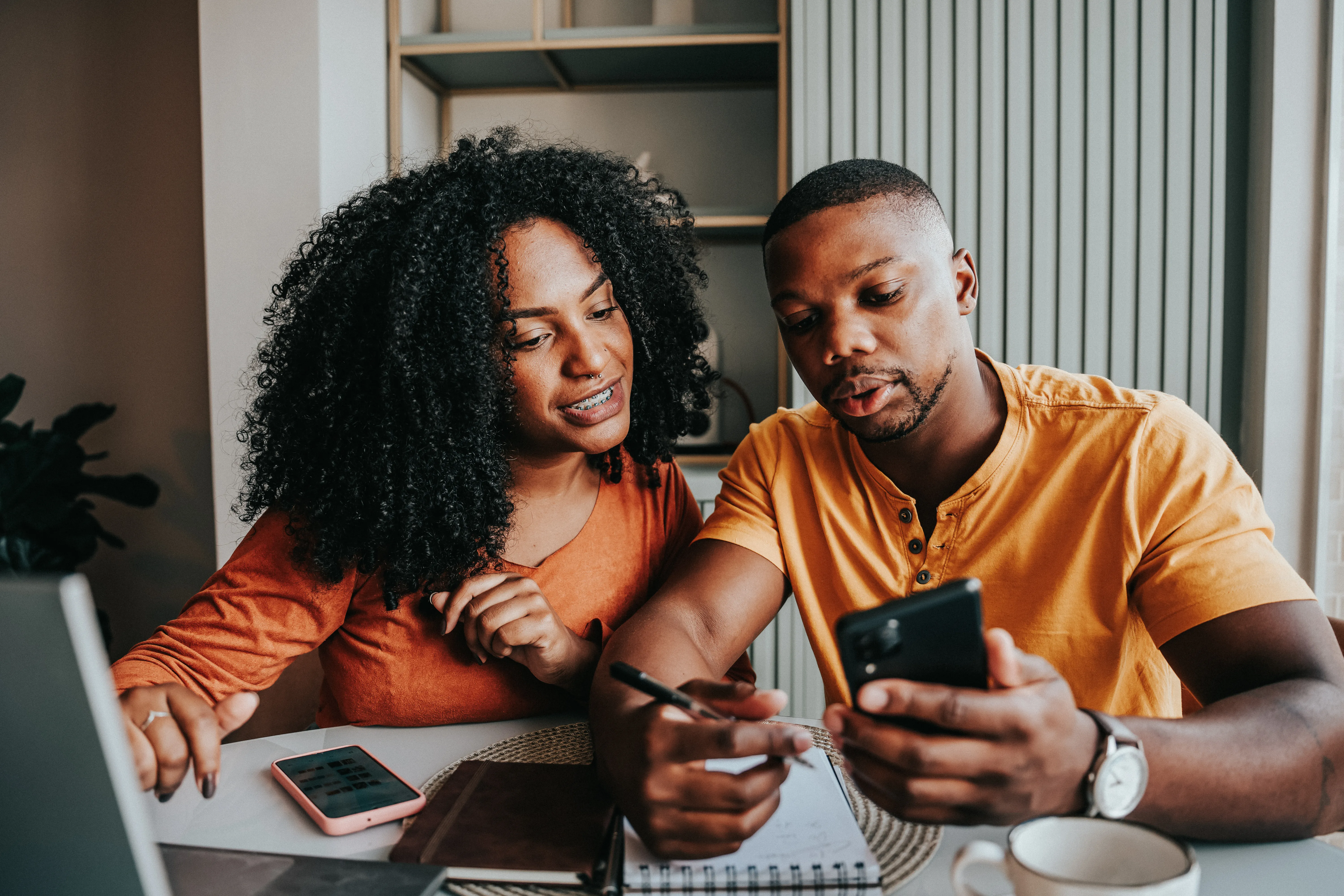 Two people sitting at a table indoors, looking at a smartphone together while one writes in a notebook 