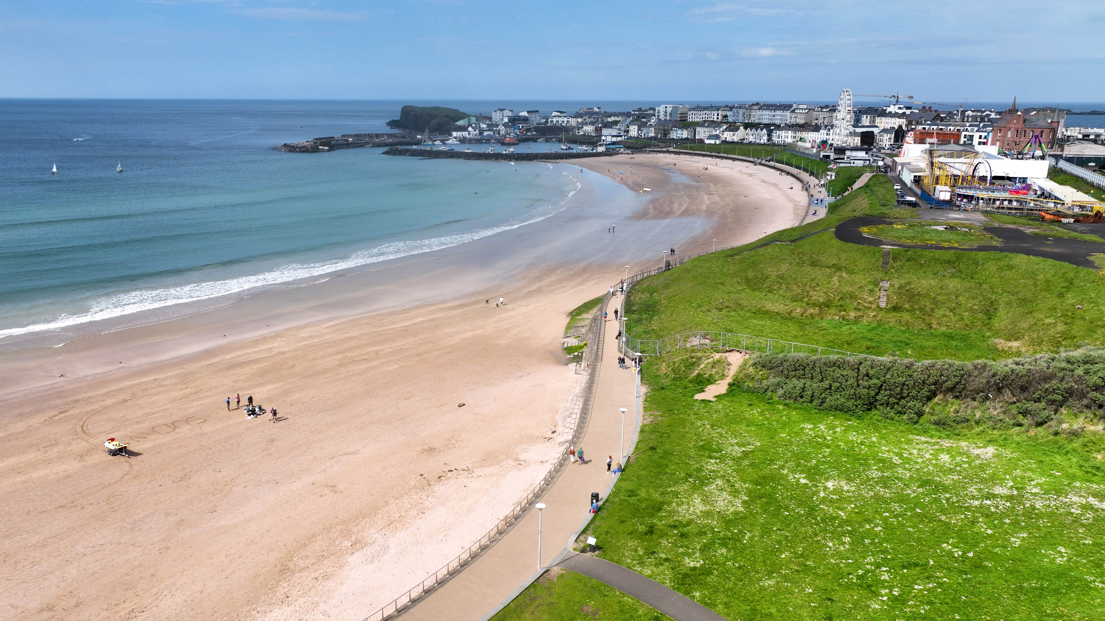 A wide sandy beach curves around the bay in Portrush, Northern Ireland, with calm blue waters and a town filled with white buildings and a Ferris wheel in the background. A promenade and grassy hillside run alongside the beach.