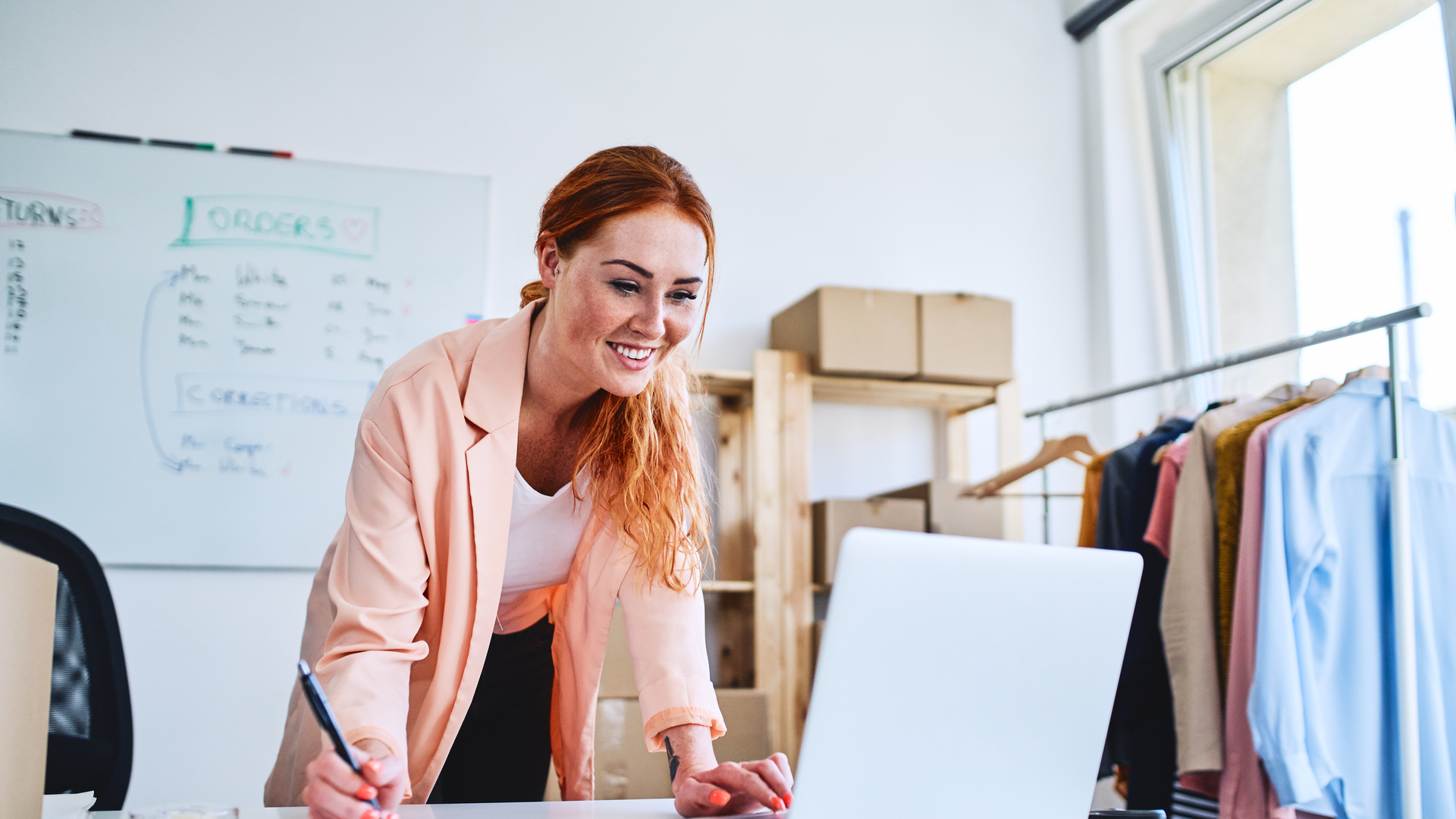 Female business owner leaning on desk in office and looking at laptop
