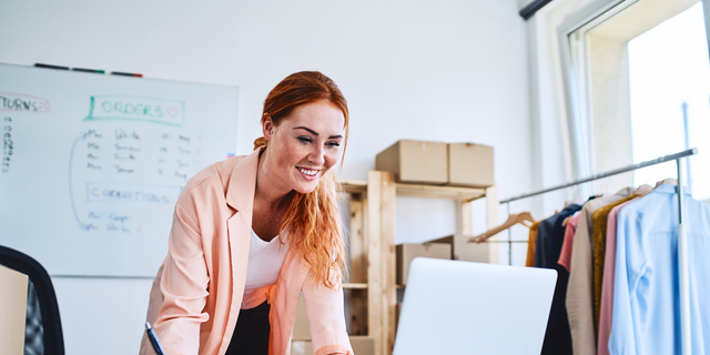 Female business owner leaning on desk in office and looking at laptop
