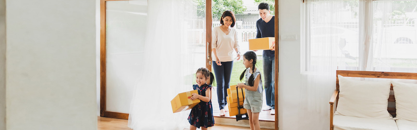 An Asian woman and man walk into a new home carrying boxes and smiling at their two young girls 