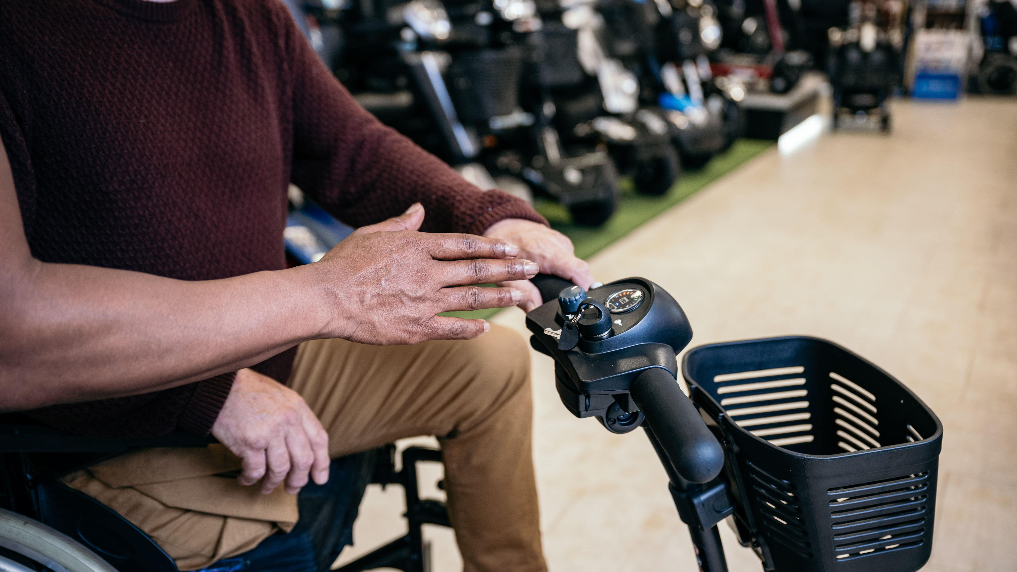 A close-up of a man’s hands operating the control panel of a mobility scooter inside a showroom, with other scooters visible in the background.