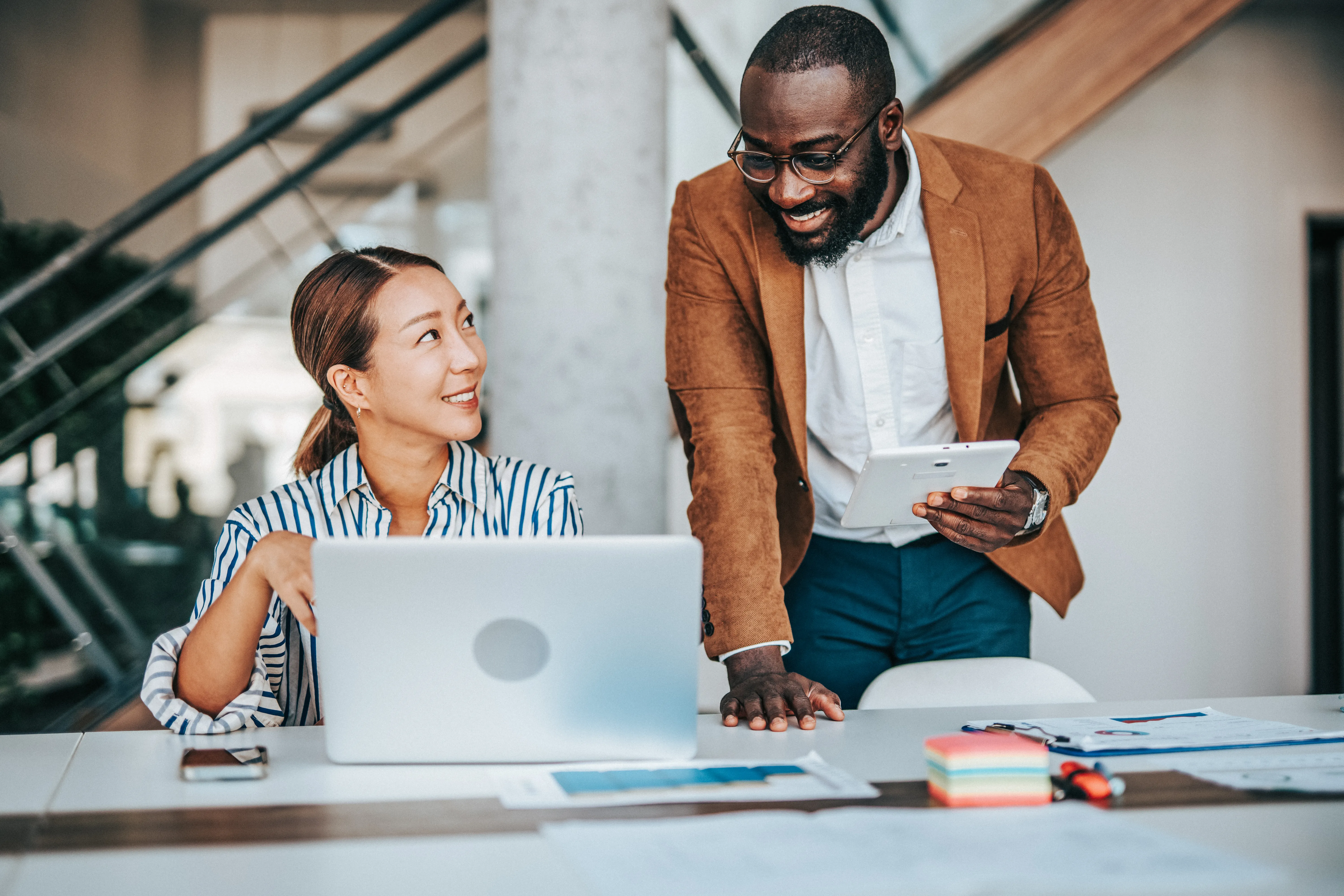 Man standing next to seated female coworker looking at laptop in office 
