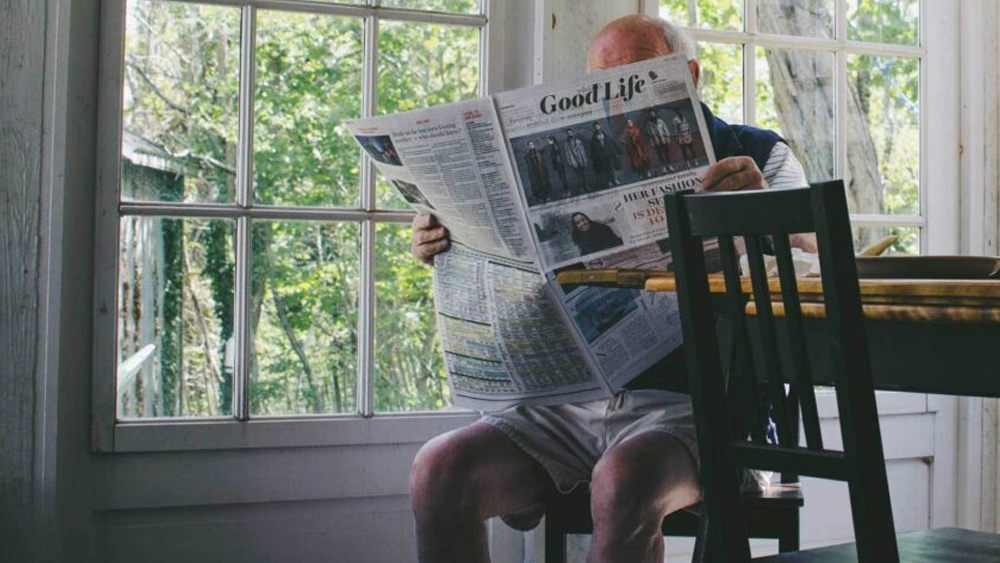 Picture of an elderly man in shorts with newspaper sitting at a dining table