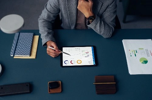 Image of a person in front of a dark green desk with a tablet, notebooks, papers, and office supplies