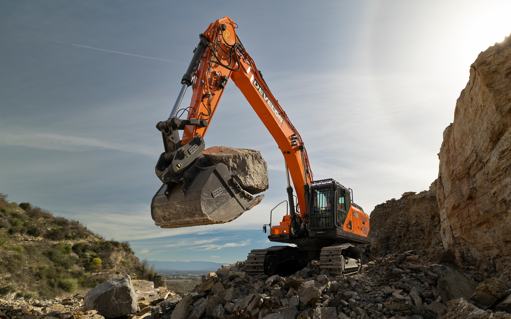 A DEVELON DX400LC-9 crawler excavator lifting a large boulder in its bucket at a rocky quarry site.