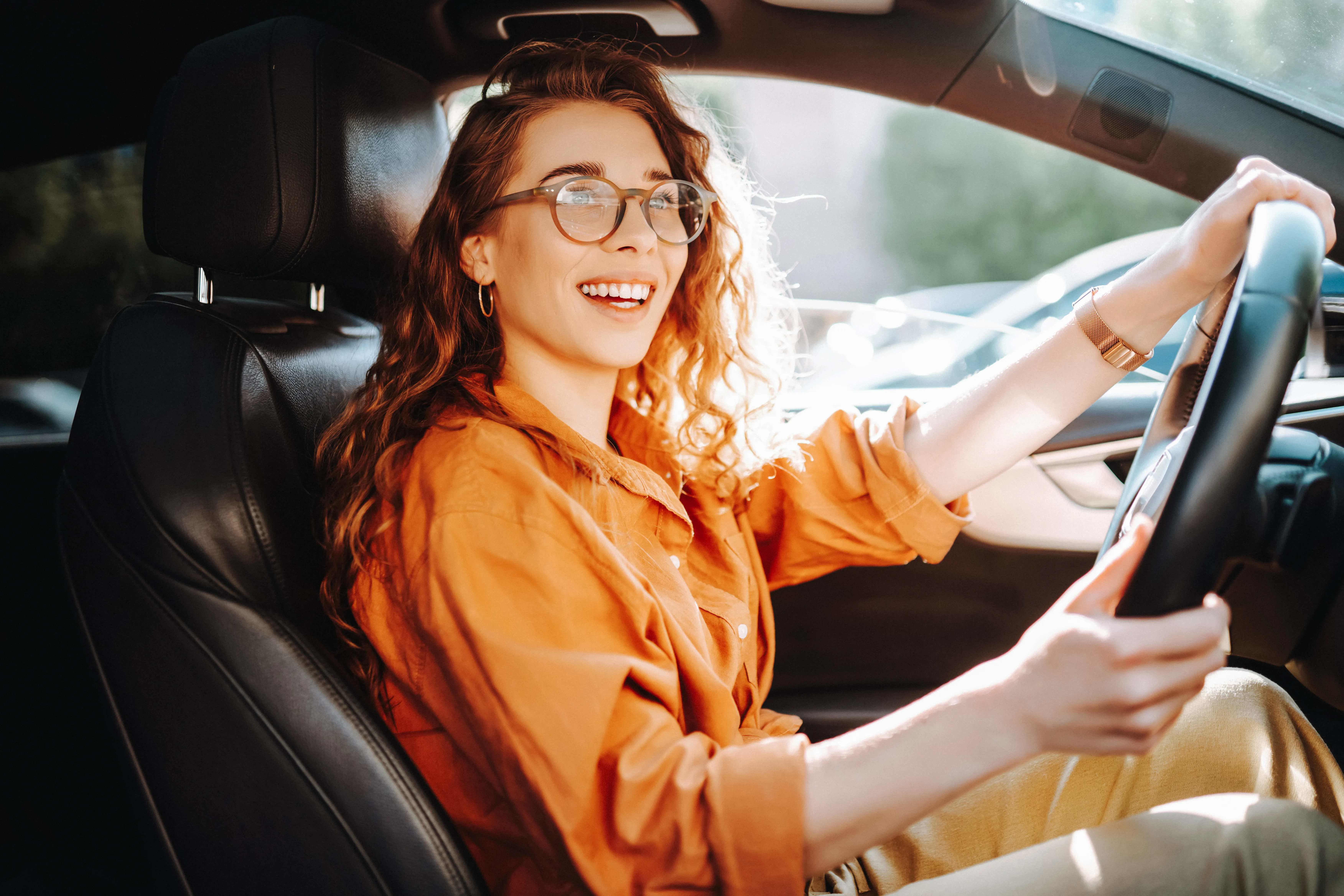 A young woman wearing glasses and brightly colored clothing is in the driver’s seat, driving a dark interior colored vehicle.  