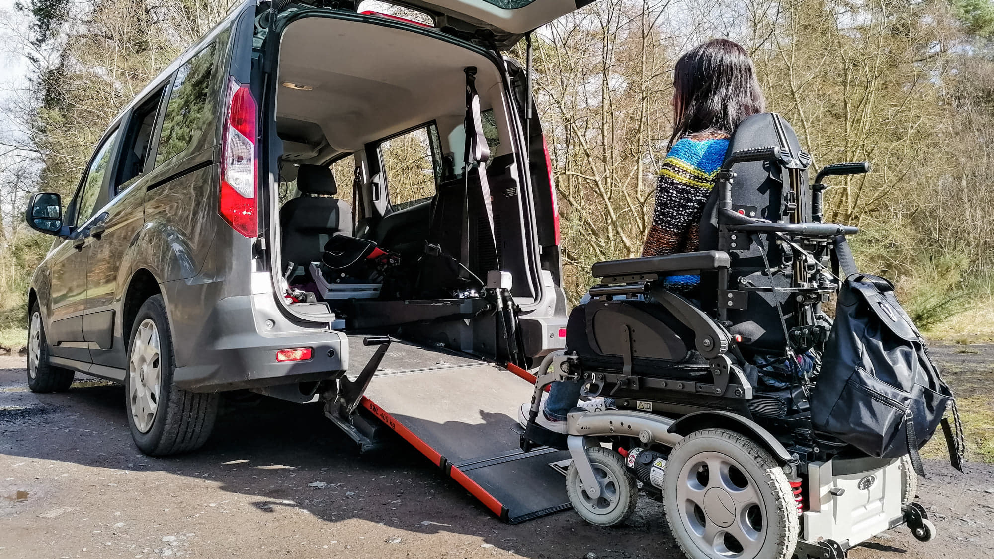 Emma Muldoon approaches a wheelchair accessible vehicle (WAV) from behind, seated in her powered wheelchair as the rear ramp is deployed in an outdoor woodland setting.