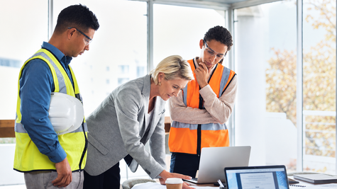 Female executive reviewing architectural blueprints and laptop data with field technicians in safety vests