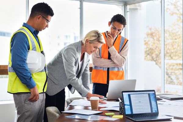 Female executive reviewing architectural blueprints and laptop data with field technicians in safety vests