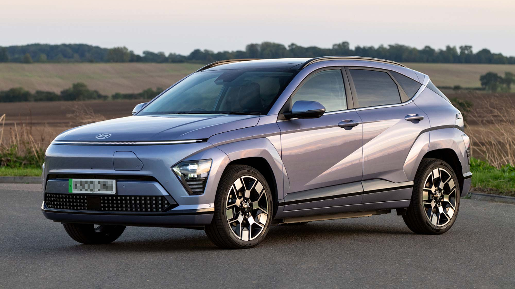 A metallic blue Hyundai Kona Electric SUV parked in a rural setting at sunset, with sharp lines and distinctive LED lighting.