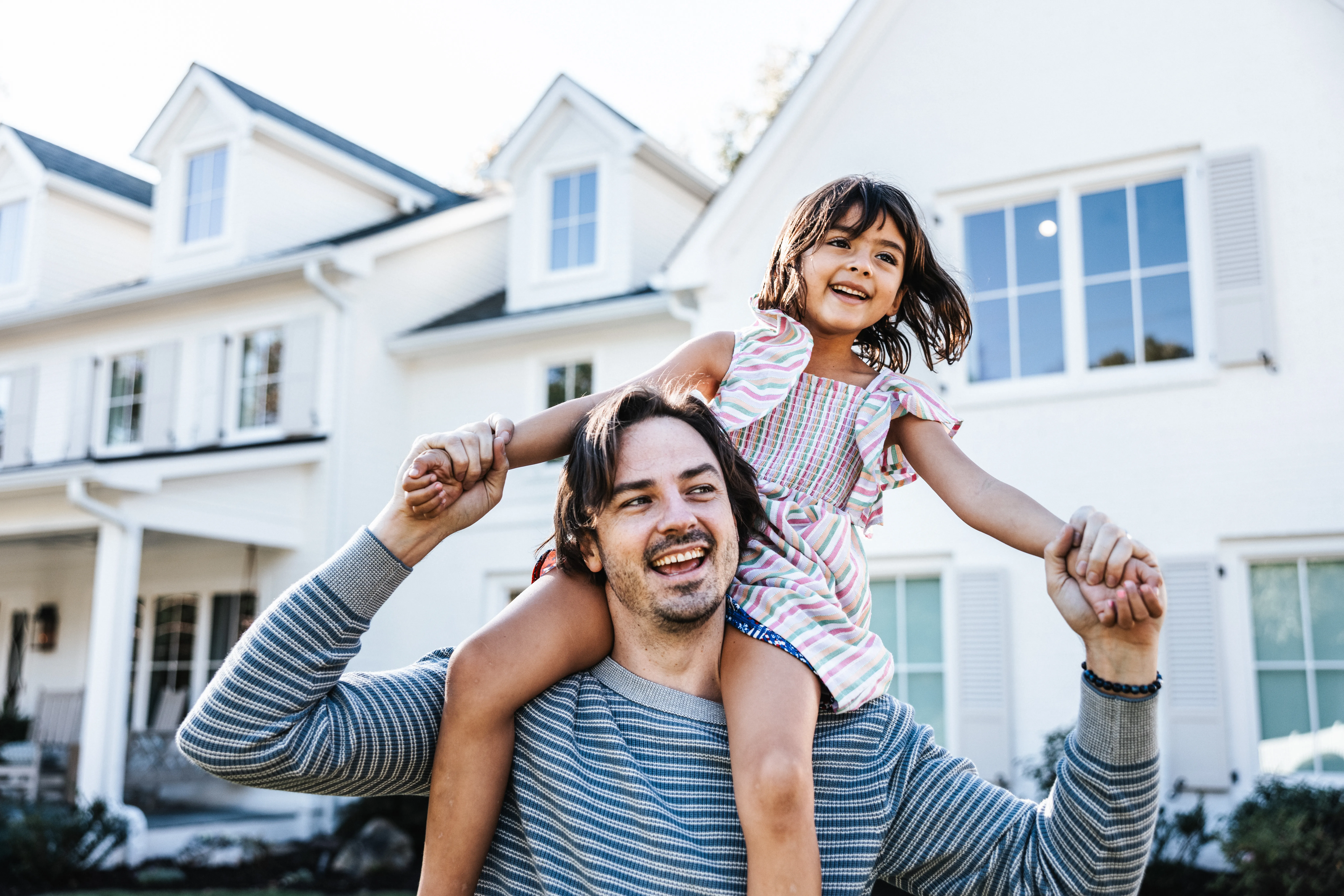 Young girl riding on her father’s shoulders in front of a white suburban home. The two are holding hands with arms extended outward. 