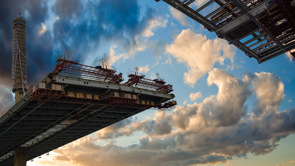 Photo of a bridge under construction taken from below with blue cloudy sky in background