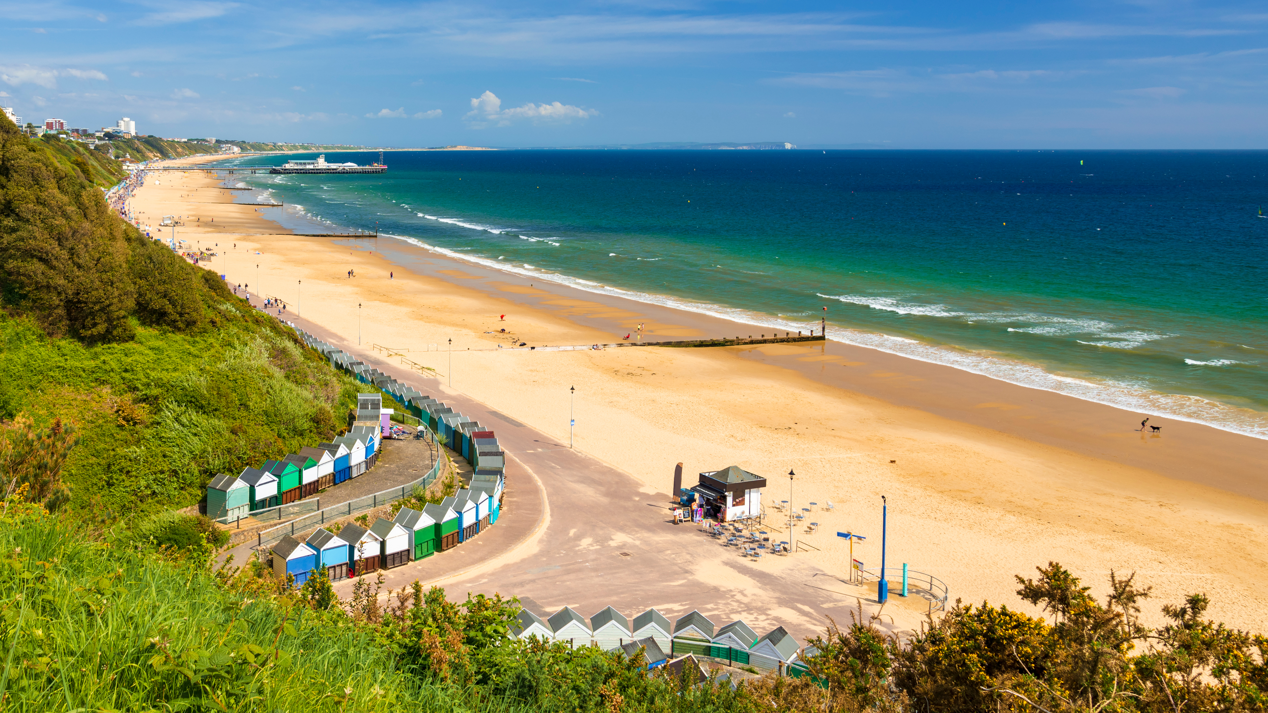 A panoramic view of Sandbanks Beach featuring soft golden sand, colourful beach huts, and a sweeping promenade. The coastline stretches toward a distant pier with deep blue water and a vibrant sky.