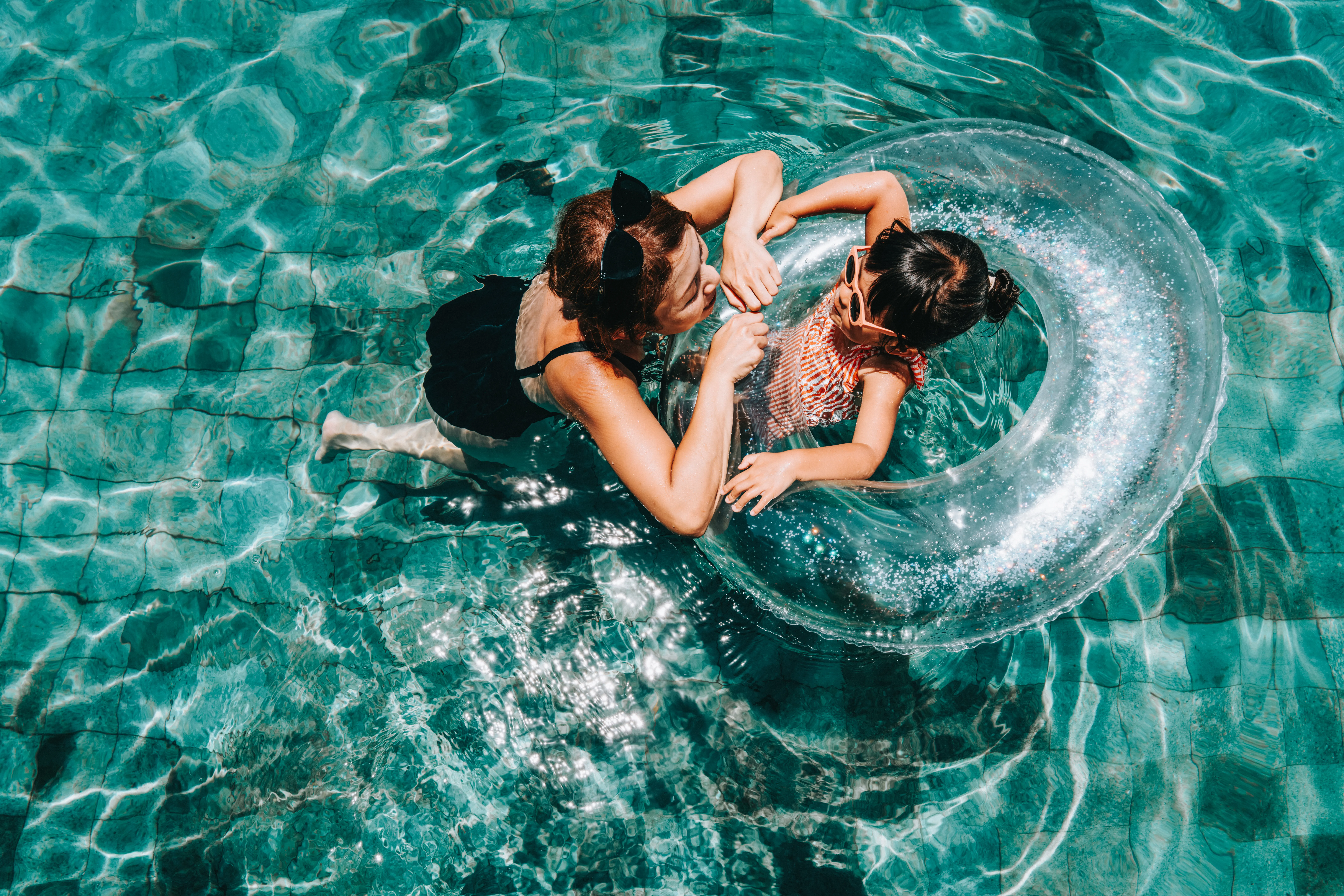 Mother floating in a pool gently guides her young daughter, who is in a water ring, through the sparkling, tiled pool. 
