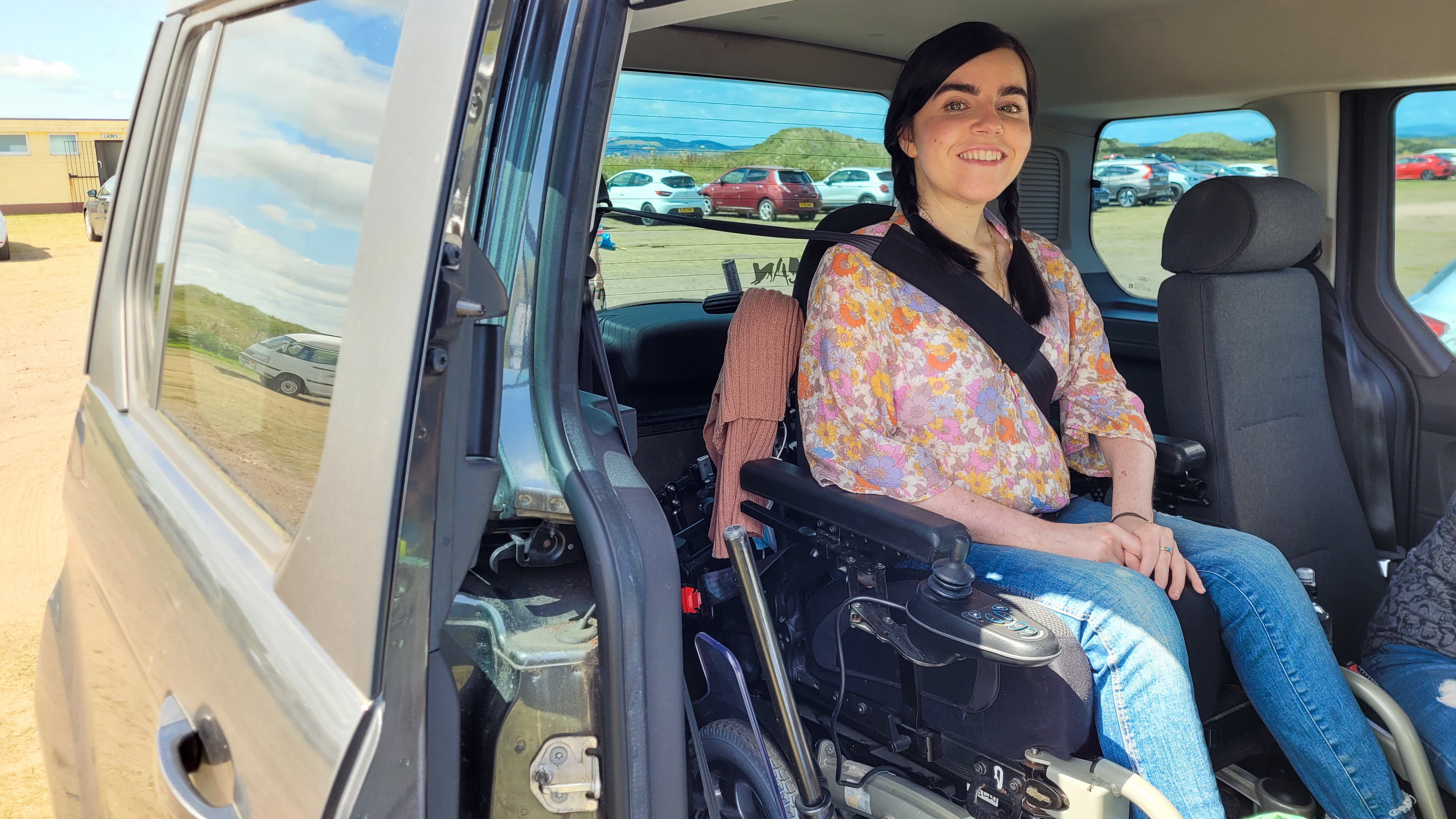 Emma Muldoon smiles from inside the back of a WAV, sitting comfortably in her powered wheelchair with the door open, parked in a coastal or rural area.
