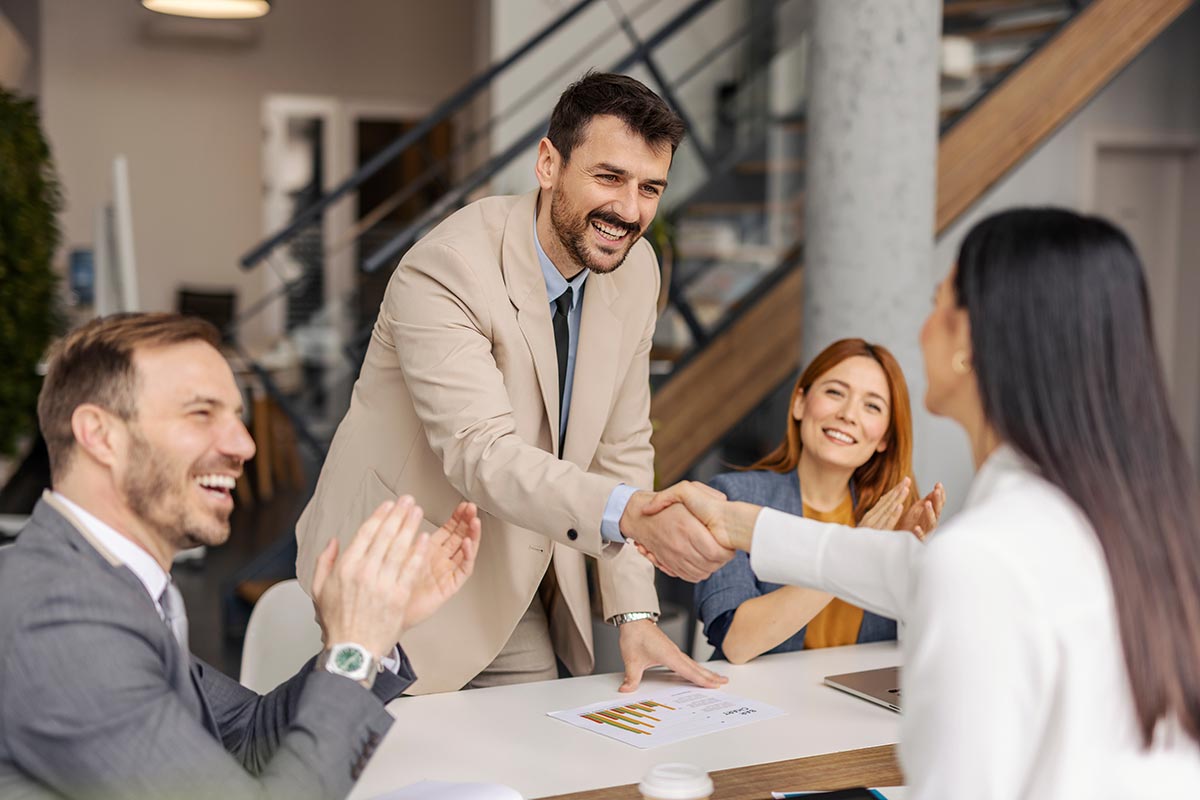 Smiling executive standing at corporate office and shaking hands with successful businesswoman while coworkers applauding.