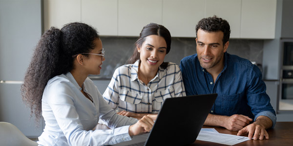 A woman sits at a table with another woman and man, she is showing them something on a laptop and they are all smiling