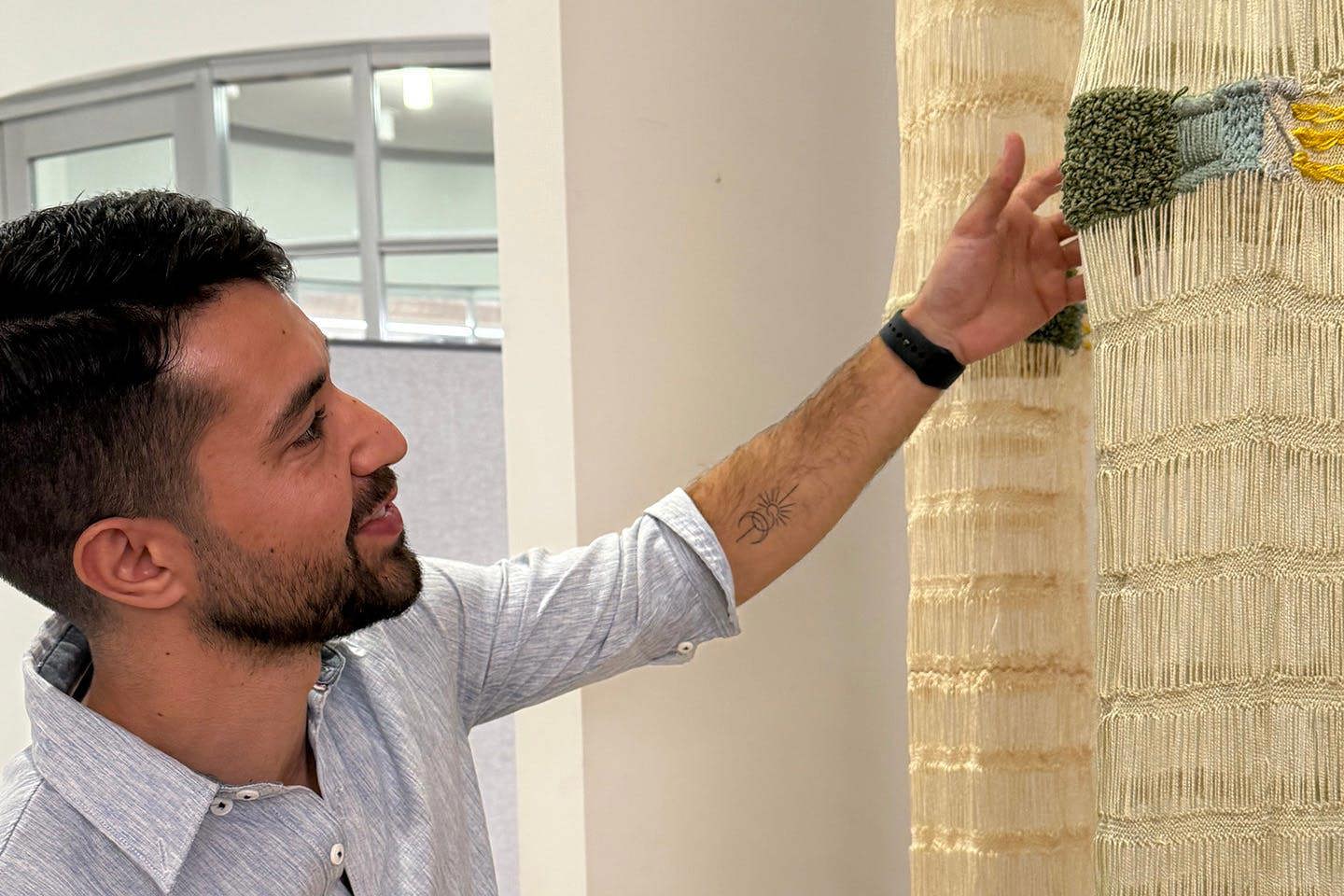 Luciano Arrorio extends a hand to closely examine a woven student artwork composed of cream-colored vertical panels accented with textured sections of green, blue, and yellow yarn.