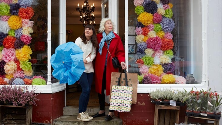 Two women walking past a shop window on a rainy day, sharing a transparent umbrella. One woman wears a red coat and carries shopping bags, while the other, in a beige coat, holds the umbrella. They are engaged in conversation, smiling, as they navigate the wet pavement