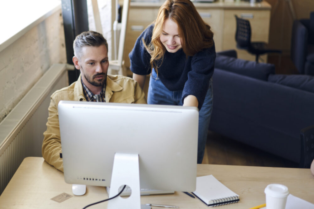 Image of a male and female worker at Desktop computer. Female is standing next to male sitting.