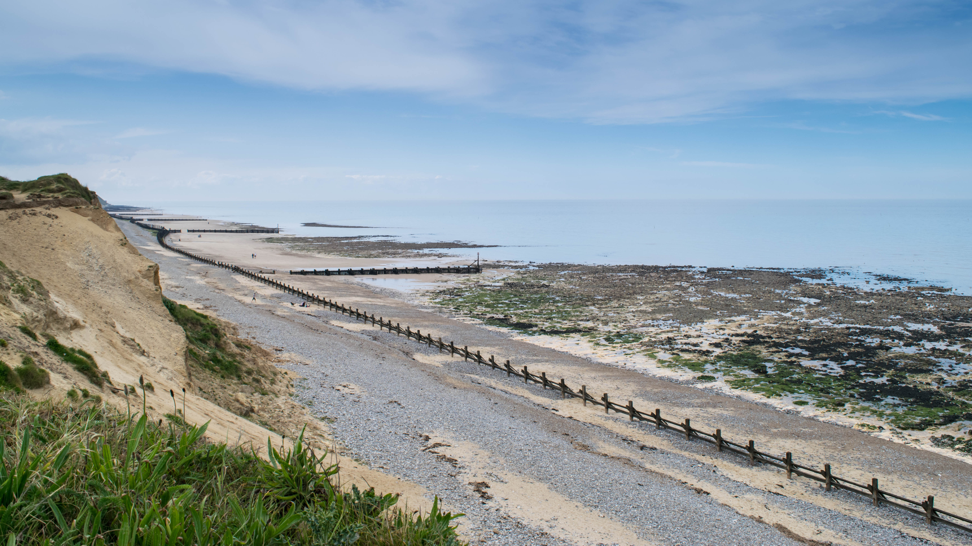 West Runton beach at low tide with a rocky shoreline and wooden groynes stretching into the sea. The sandy cliffside and pebbled beach lead into calm waters beneath a partly cloudy sky.