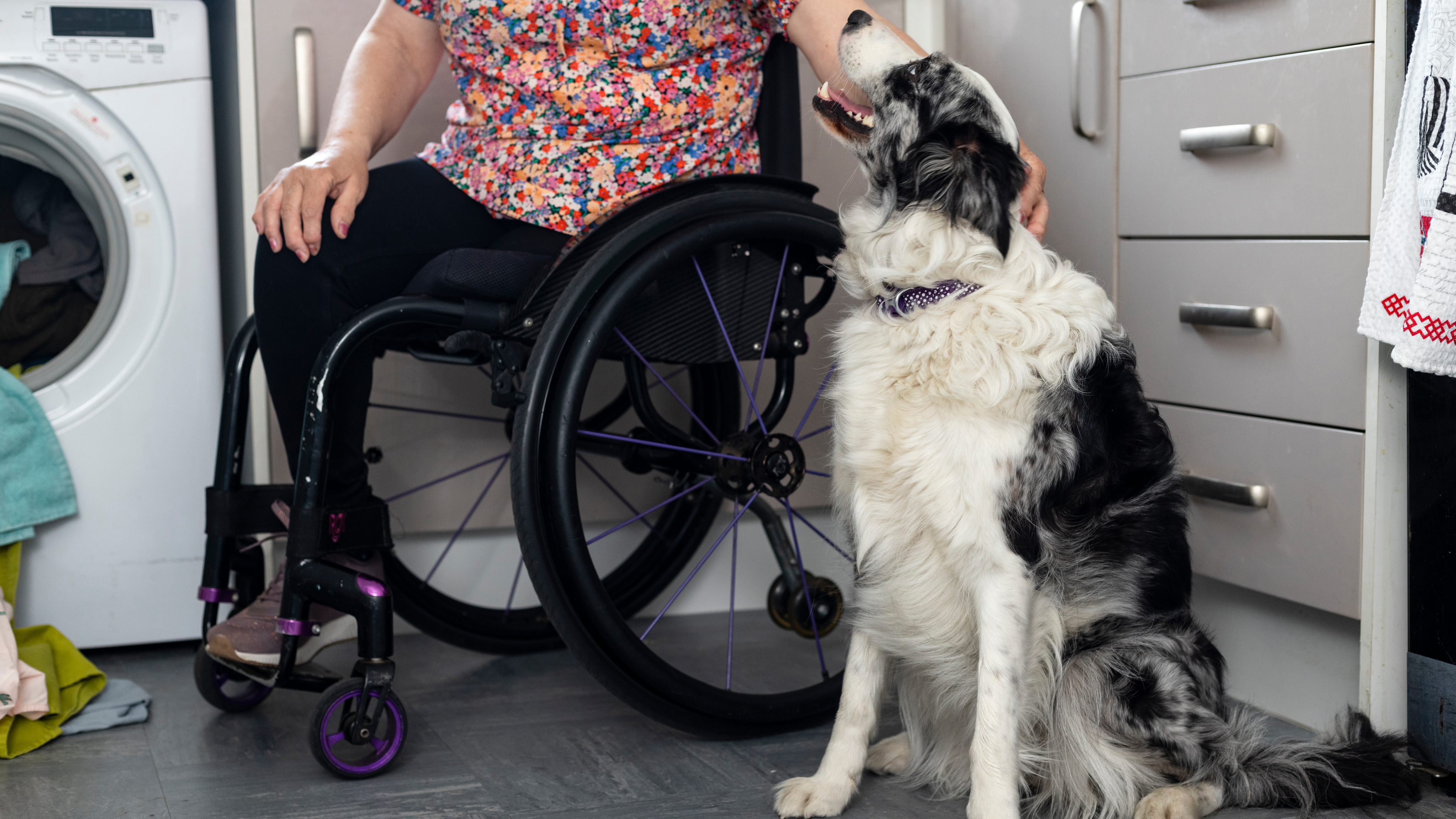 A person in a wheelchair, wearing a colorful floral top, gently strokes the head of a large black and white assistance dog sitting beside them in a kitchen. The dog looks up affectionately at the person. Nearby, a washing machine is partially visible with laundry on the floor, adding to the cozy, lived-in feel of the space.