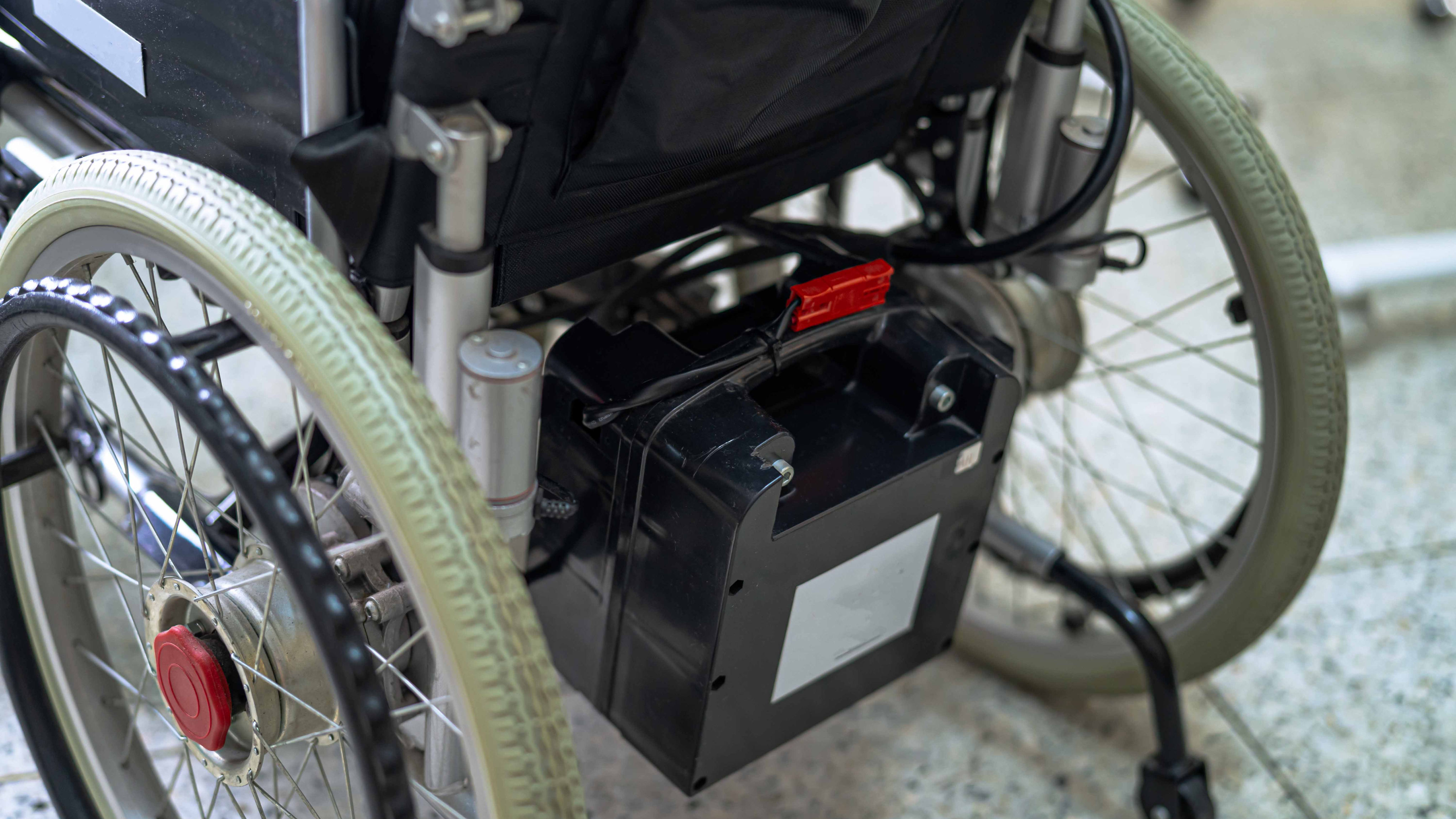 "Close-up of the rear section of a powered wheelchair, focusing on the black battery pack attached beneath the seat. The wheelchair has sturdy wheels with light-coloured tyres, metal spokes, and a red central hub. The background features a tiled floor and blurred indoor surroundings.
