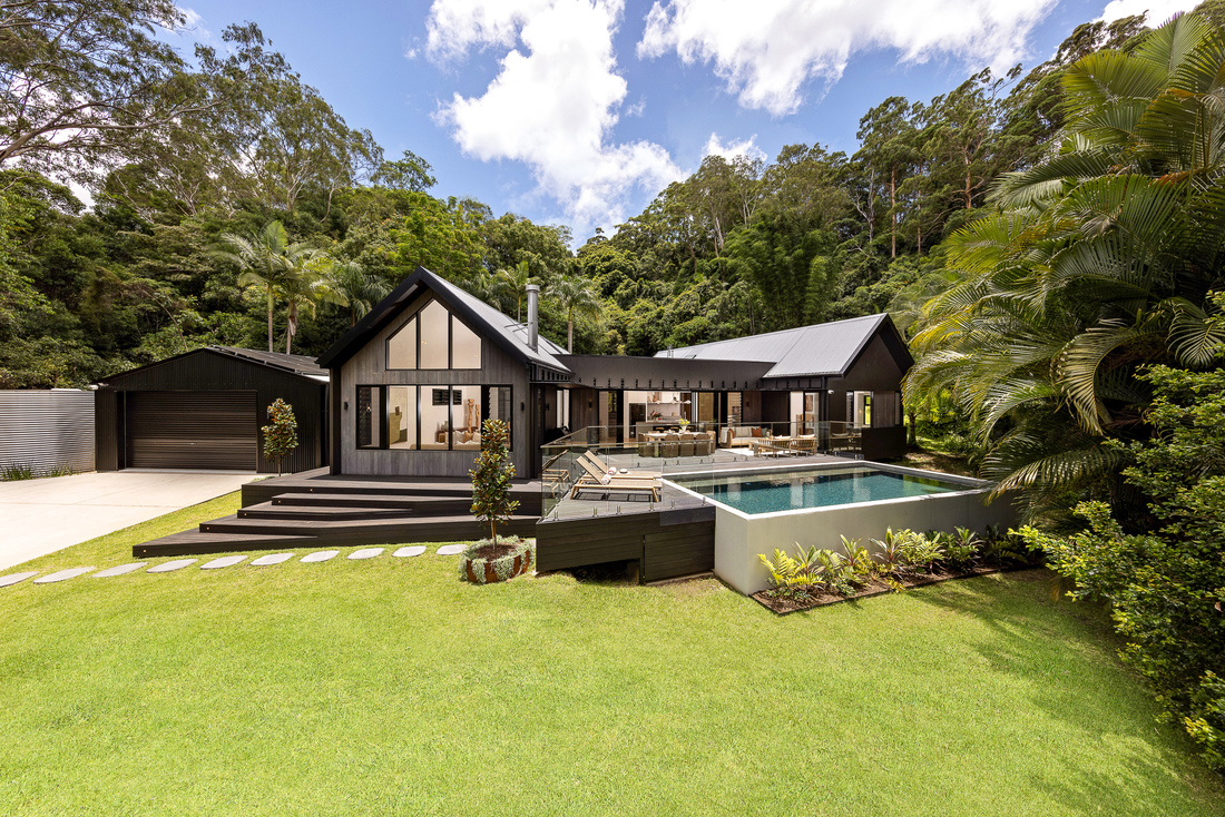 yourtown Prize Home Draw 545. External image from the front of a single-level pavilion-style house with black cladding. There is an alfresco area at the front with a pool. A garage and driveway is on the left hand side.