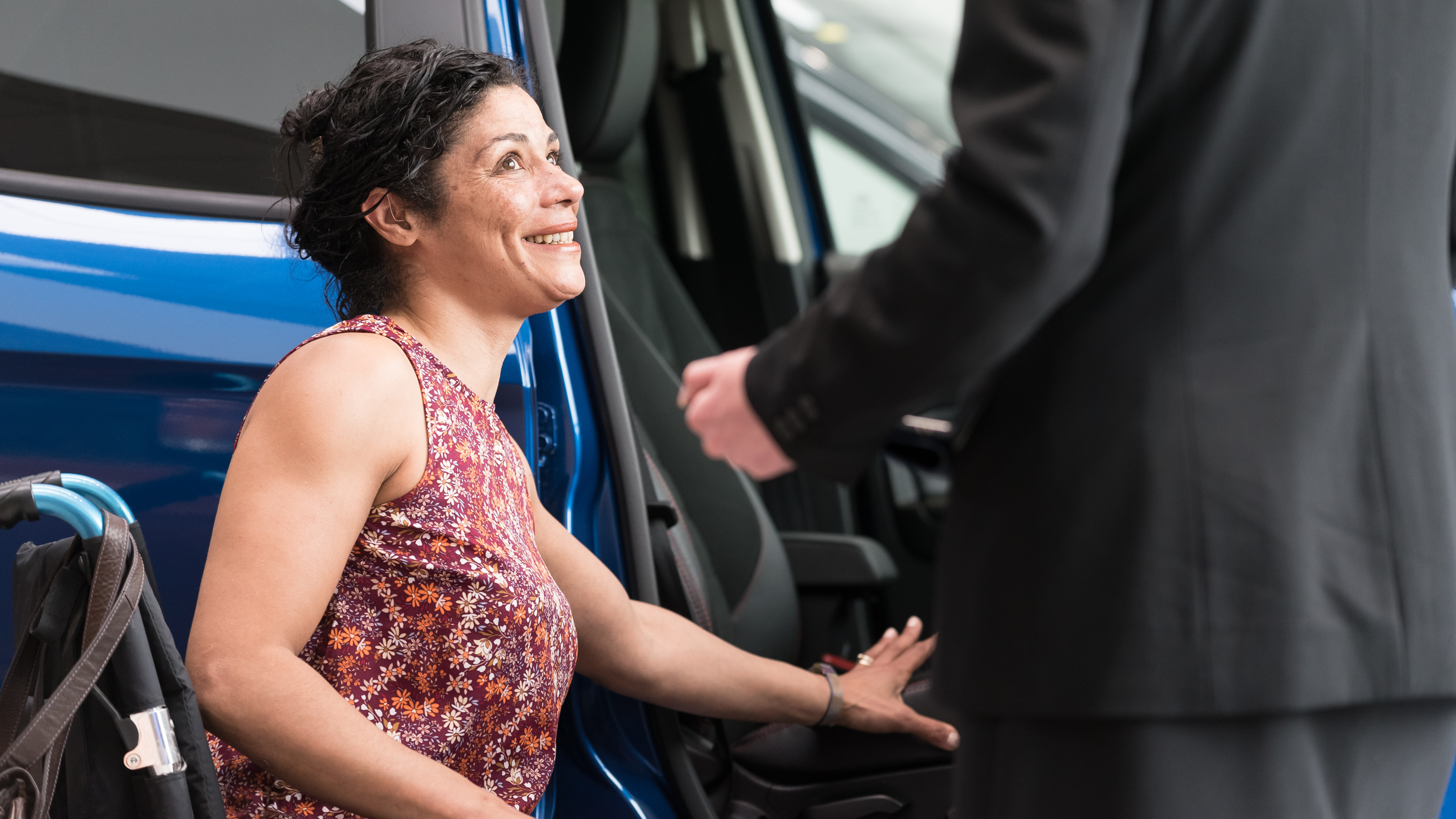 A woman using a wheelchair smiles as she transfers into the front seat of a blue car. A person standing nearby, dressed in a suit, is assisting her, suggesting a supportive or customer service interaction at a dealership or mobility vehicle provider.