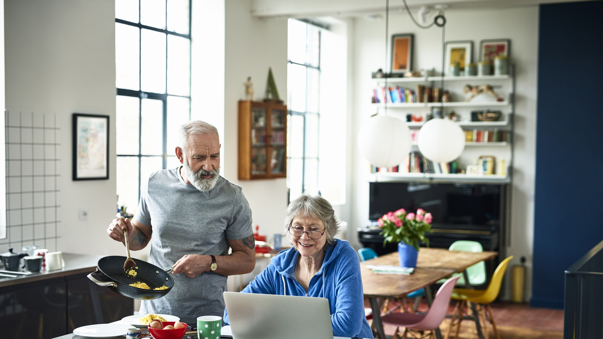 Senior woman using laptop and smiling as man serves breakfast