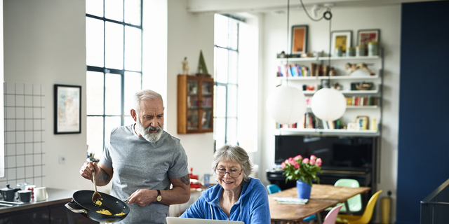 Senior woman using laptop and smiling as man serves breakfast