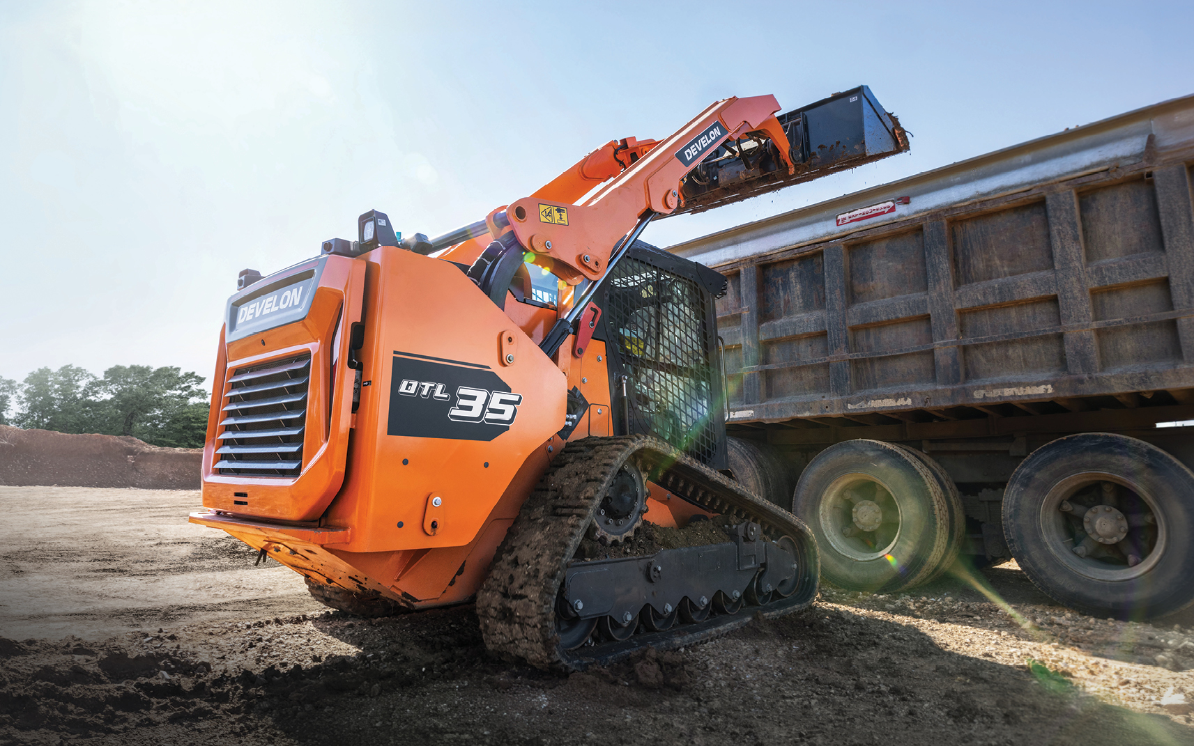 DEVELON DTL35 compact track loader lifting a bucket of dirt into a high-sided dump truck.