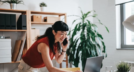 Professional Asian woman on phone leaning over desk typing on laptop in small office 